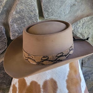May include: A tan cowboy hat with a snakeskin band. The hat has a wide brim and a rounded crown. The hat is displayed on a cowhide surface.