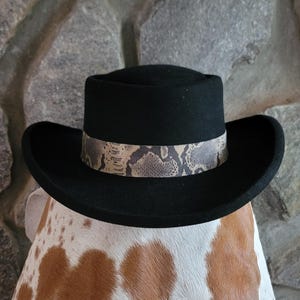 May include: A black felt hat with a flat crown and a wide, curved brim. The hat features a decorative band around the base of the crown, displaying a textured, light brown and grey snakeskin pattern that contrasts with the solid black material.