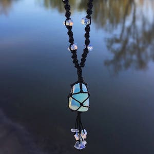 Puede incluir: Un collar con un colgante de piedra de ópalo azul y blanco, cordón de macramé negro y cuentas de ópalo blanco. El collar está colgado sobre un fondo borroso de una masa de agua.