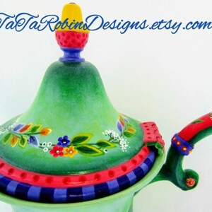 May include: A green ceramic teapot with a red and yellow lid and a red and white handle. The teapot is decorated with painted flowers and leaves.