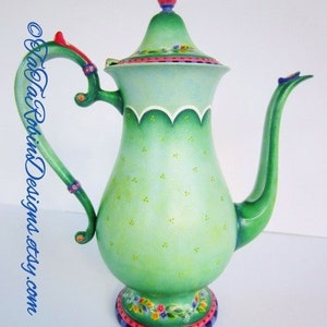 May include: A green ceramic teapot with a floral design and a yellow bird on the lid. The teapot has a scalloped edge and a green and blue base.