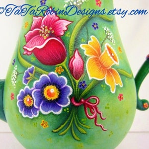 May include: A green ceramic teapot with a floral design. The teapot is painted with pink, yellow, blue, and white flowers. The teapot has a handle and a spout. The text "TaTa Robins Designs.etsy.com" is painted on the side of the teapot.