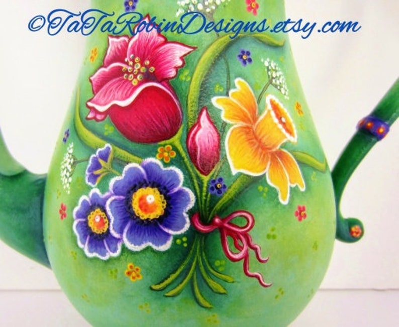 May include: A green ceramic teapot with a floral design. The teapot is painted with pink, yellow, blue, and white flowers. The teapot has a handle and a spout. The text "TaTa Robins Designs.etsy.com" is painted on the side of the teapot.