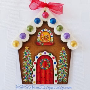 Peut inclure: Une d&eacute;coration de No&euml;l en forme de maison en pain d'&eacute;pices avec une porte rouge, du gla&ccedil;age blanc et un personnage en pain d'&eacute;pices regardant par la fen&ecirc;tre. La d&eacute;coration est orn&eacute;e de points de bonbons color&eacute;s et d'&eacute;toiles.