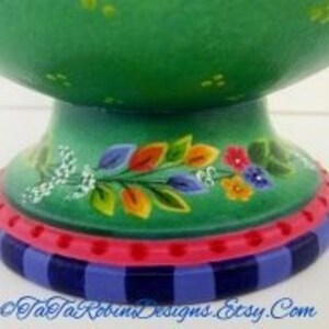 May include: Green ceramic bowl with a floral design on the base. The base has a blue and white striped pattern with a pink band.
