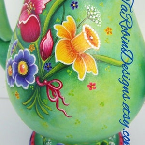 May include: A green ceramic pitcher with a painted floral design. The design features pink, yellow, and blue flowers, green leaves, and a pink ribbon. The pitcher has a white base with a pink, blue, and black trim.