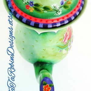 May include: A green teapot with a floral design and a matching lid. The teapot has a scalloped edge and a handle with a purple and orange flower detail. The lid has a red and purple trim.