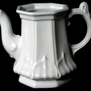 May include: A white porcelain teapot with a octagonal body and a decorative, ornate handle. The teapot has a smooth, glossy finish and a delicate, floral design.