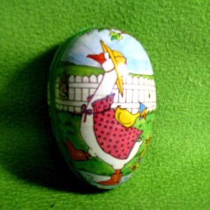 May include: A white ceramic egg-shaped container with a colorful illustration of a goose wearing a red and white polka dot dress and a yellow chick. The goose is standing in a green grassy area with a white picket fence in the background.