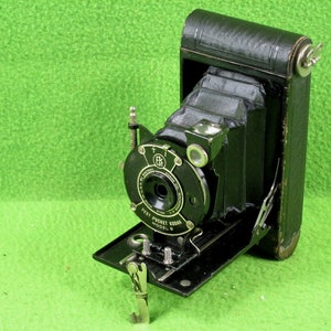 May include: A black folding camera with a leather exterior. The camera has a lens with a silver ring and the text "Vest Pocket Kodak Model 8" printed on the front.