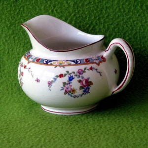 May include: A white ceramic creamer with a red, blue, and gold floral design. The creamer has a handle and a rounded body.