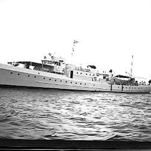 May include: A black and white photo of a large, white ship with a flag flying from the mast. The ship is in the water and there is a smaller boat behind it.
