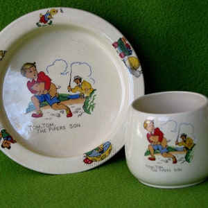 May include: A set of vintage children's dishes featuring a cartoon illustration of a boy with a pig. The plate and mug are both white with a yellow rim and feature the text "Tom. Tom. The Pipers Son".