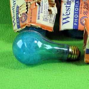 May include: A vintage blue-green light bulb with a gold base, resting on a green surface. Partially visible are several orange and white Westinghouse Photo Flash boxes, with text and logos. The boxes are torn and aged.