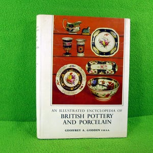 May include: A book titled "An Illustrated Encyclopedia of British Pottery and Porcelain" by Geoffrey A. Godden F.R.S.A. The cover features a colorful illustration of various pieces of pottery and porcelain, including plates, bowls, and a teapot.