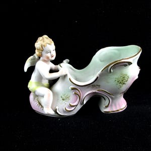 May include: A porcelain shoe-shaped figurine with a cherub figure. The shoe is primarily white with pink and gold accents, and the cherub has blonde hair and wings. The figurine is likely a decorative item.