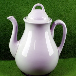 May include: A white ceramic teapot with a lavender glaze and a lid. The teapot has a rounded body and a spout on the left side. The handle is on the right side.