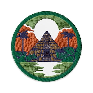 Può includere: Una toppa circolare ricamata con un bordo verde e uno sfondo bianco. La toppa presenta una piramide marrone con una cima blu, circondata da vegetazione verde e montagne arancioni. Il sole splende sopra la piramide.