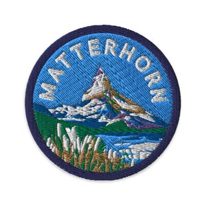 Può includere: Toppa rotonda ricamata raffigurante il Cervino, con la scritta "MATTERHORN" in bianco. La toppa presenta uno sfondo blu e una scena di montagna in verde, bianco, viola e marrone. Ha un bordo blu scuro.