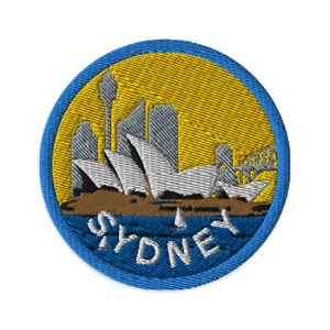 Op de afbeelding: Een blauwe en gele geborduurde patch met een wit silhouet van het Sydney Opera House en de Sydney Harbour Bridge. De tekst "SYDNEY" staat in een blauwe boog rond de onderkant van de patch.