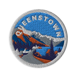 May include: A round, embroidered patch featuring a blue sky, white mountains, and a forest scene. The text "Queenstown" is stitched in white around the edge of the patch.