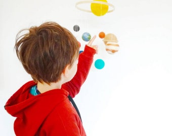 May include: A child in a red hoodie points at a colorful solar system mobile. The mobile features a yellow sun with a ring, and planets in various colors, including blue, orange, and brown. The planets are suspended from a white string.
