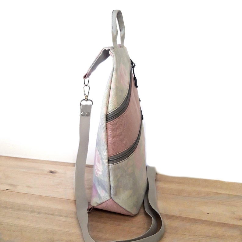 May include: A floral-patterned backpack with a pink stripe down the front. The bag has a gray handle and shoulder strap, and a black zipper. The bag is made of a soft, textured material.