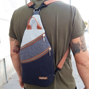 May include: A multi-tone sling bag with a gray, denim blue, and navy blue color scheme. The bag features brown zipper accents and straps, with the text "myPeri" on the bottom. The bag is worn over the shoulder.