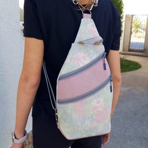 May include: A floral print sling bag with a pink and gray color scheme. The bag features a pink stripe across the front with two gray zippers. The bag is worn over the shoulder.