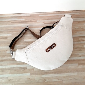 May include: Cream-colored canvas fanny pack with a brown leather strap and zipper closure. The bag has a curved shape and a small brown leather tag that reads "myPeri". The bag is on a wooden surface.