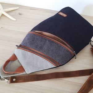 May include: A navy blue and gray sling bag with a brown leather strap and zipper accents. The bag features a brown leather patch with the brand name. The bag is made of durable fabric and has a modern design.