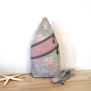 May include: A floral-patterned sling bag with a pink stripe and two zippered pockets. The bag features a gray adjustable strap and a small handle at the top. The floral pattern includes pink roses on a gray background.