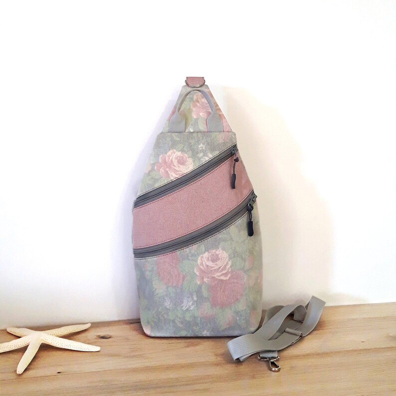 May include: A floral-patterned sling bag with a pink stripe and two zippered pockets. The bag features a gray adjustable strap and a small handle at the top. The floral pattern includes pink roses on a gray background.