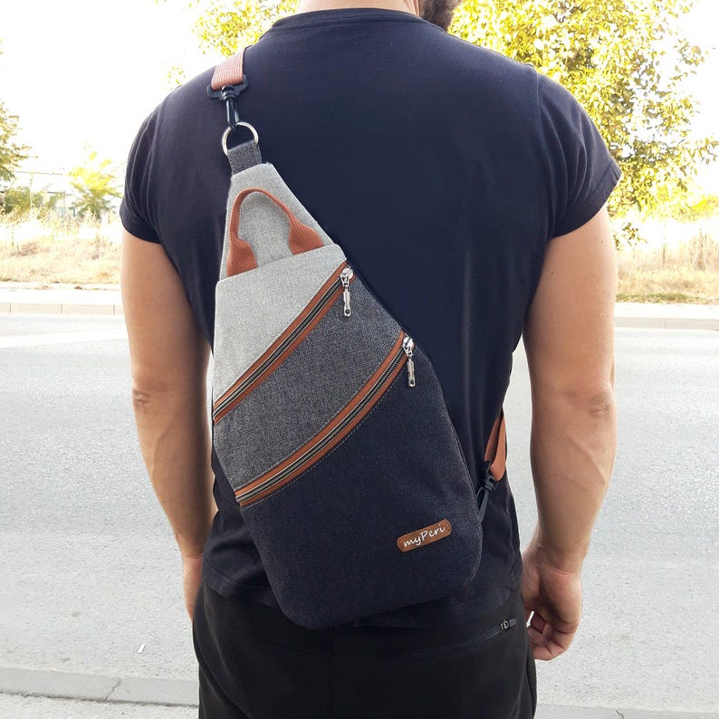 May include: A multi-tone sling bag with a gray, denim, and navy blue color scheme. The bag features brown leather accents, a top handle, and two zippered compartments. The bag is worn over the shoulder and has the text "myPeru" on the bottom.
