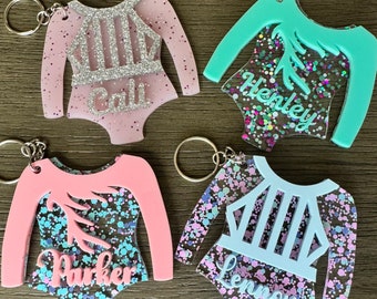 May include: Four colorful keychains shaped like leotards with glitter accents. Each keychain has a different name on it: Cali, Henley, Parker, and Lennox.