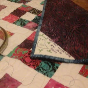 May include: A white quilt with a patchwork design featuring various colors and patterns. A purple fabric with a blue trim is folded over the quilt. The fabric has a white embroidered bird with the text "Happy Winter Love by Jenn 2017".