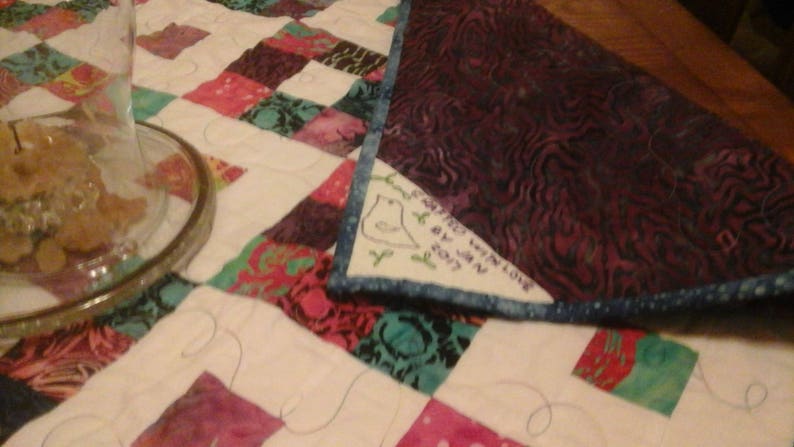 May include: A white quilt with a patchwork design featuring various colors and patterns. A purple fabric with a blue trim is folded over the quilt. The fabric has a white embroidered bird with the text "Happy Winter Love by Jenn 2017".