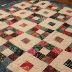 May include: A white quilt with a blue border. The quilt is made of squares of different fabrics, each with a unique pattern. The fabrics are mostly shades of blue, green, and purple.