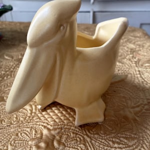 Könnte beinhalten: Ein Keramik-Pflanzgefäß in Form eines Pelikans, mit hellgelber Glasur. Der Vogel hat einen langen Schnabel und eine weite Öffnung zum Bepflanzen. Ein dekorativer Artikel.