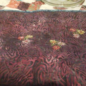 May include: A close-up of a dark purple quilted fabric with a zebra-like pattern. The fabric is decorated with embroidered flowers in shades of pink, yellow, and green.
