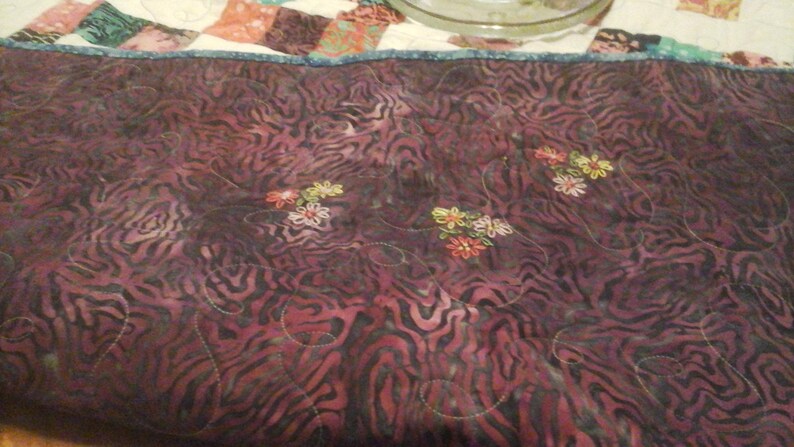 May include: A close-up of a dark purple quilted fabric with a zebra-like pattern. The fabric is decorated with embroidered flowers in shades of pink, yellow, and green.