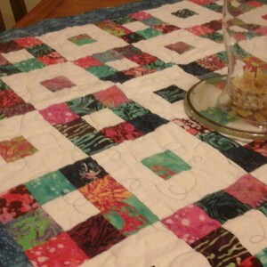 May include: A white table runner with a blue border and a patchwork design of colorful squares. The runner is made of fabric with a floral pattern. A clear glass vase is sitting on the runner.