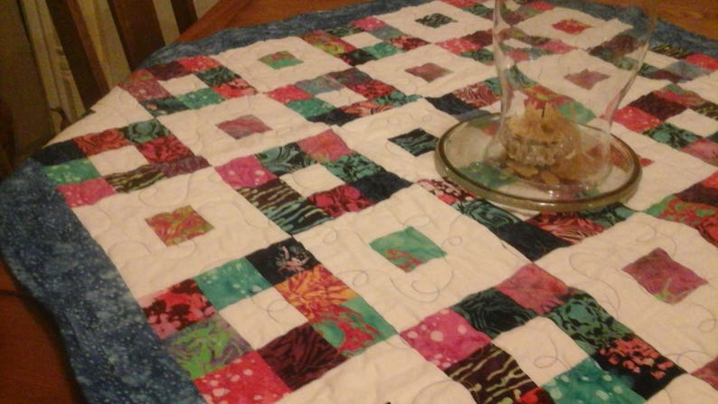 May include: A white table runner with a blue border and a patchwork design of colorful squares. The runner is made of fabric with a floral pattern. A clear glass vase is sitting on the runner.