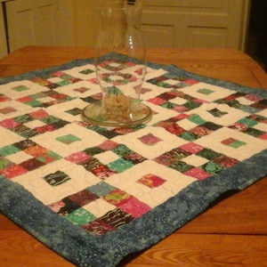 May include: A patchwork quilt table runner with a blue border and a white background. The runner is made of various colorful fabrics in shades of pink, green, and brown. A clear glass vase sits in the center of the runner.