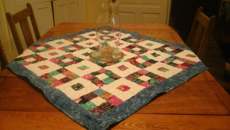 May include: A patchwork quilt table runner with a blue border and a white background. The runner is made of various colorful fabrics in shades of pink, green, and brown. A clear glass vase sits in the center of the runner.