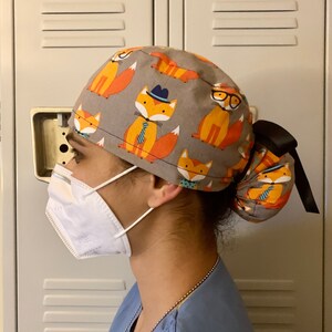 Puede incluir: Una persona lleva una gorra de quir&oacute;fano gris y naranja con estampado de zorro. La gorra tiene una cinta negra y est&aacute; atada en un mo&ntilde;o en la parte posterior. La persona tambi&eacute;n lleva una mascarilla facial blanca y una bata de quir&oacute;fano azul.