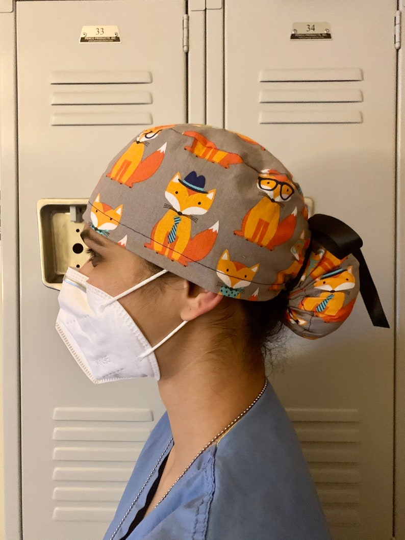 Puede incluir: Una persona lleva una gorra de quir&oacute;fano gris y naranja con estampado de zorro. La gorra tiene una cinta negra y est&aacute; atada en un mo&ntilde;o en la parte posterior. La persona tambi&eacute;n lleva una mascarilla facial blanca y una bata de quir&oacute;fano azul.