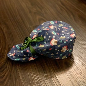 Puede incluir: Un gorro de quir&oacute;fano azul y verde con estampado y una cinta verde atada alrededor. El gorro tiene una variedad de peque&ntilde;as ilustraciones con tem&aacute;tica de perros, incluyendo huellas de patas, huesos y casetas para perros.