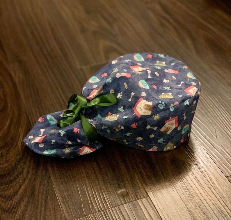 Puede incluir: Un gorro de quir&oacute;fano azul y verde con estampado y una cinta verde atada alrededor. El gorro tiene una variedad de peque&ntilde;as ilustraciones con tem&aacute;tica de perros, incluyendo huellas de patas, huesos y casetas para perros.