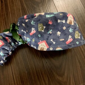 Puede incluir: Un gorro de quir&oacute;fano azul con un estampado de tem&aacute;tica canina que incluye huellas de patas, huesos, casetas para perros y bolsas de comida para perros. El gorro tiene un lazo verde.